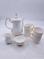 Front view of entire tea set including tea pot, four cups, creamer, and sugar bowl, all white porcelain with gold trim.
