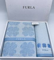 Box containing two sky blue Furla towels arranged flat showing floral pattern and branding.
