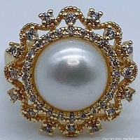 Front view showing the large central white semi-round natural pearl with scalloped decorative setting embedded with small clear stones on an 18K gold-plated band.