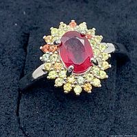 Top view of ring showing oval red central ruby and surrounding multicolor sapphire halo on black background.
