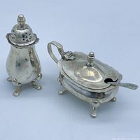 Photo showing sterling silver condiment shaker and mustard pot with spoon side by side on plain background