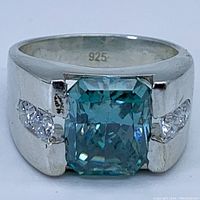Front close-up showing rectangular blue moissanite center stone and surrounding smaller clear stones on sterling silver band with 925 mark inside.