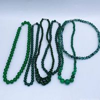 Top-down image of six green stone bead necklaces laid on a white surface, showing bead colors, sizes, and metallic clasps.
