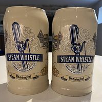 Pair of cream-colored ceramic Steam Whistle Oktoberfest beer steins with logos and decorative Oktoberfest artwork.