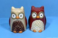 Front view of two ceramic owl shakers, one cream and brown, the other red and white, with detailed eyes and beaks.