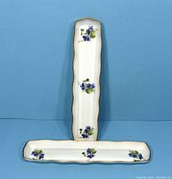 One vertical dresser tray with scalloped edges, white porcelain with three purple flower groupings and gold edge trim, placed upright against a blue background with two horizontal trays