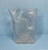 Front side of a vintage clear glass pitcher showing embossed grapevine clusters and handle.