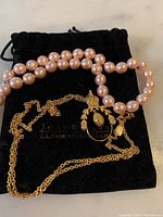 Pink pearl necklace and gold tone chain with pendant displayed on a black pouch.