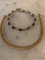 Two bracelets displayed on marble surface, one silver with red stones, one gold with clear stones