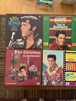 Four Elvis Presley vinyl LP records arranged on floor showing cover art: Elvis Love Songs, Elvis Fun In Acapulco, Elvis Blue Christmas, Elvis Blue Hawaii.