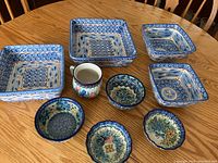 Top view of nine ceramic serving pieces arranged on a wooden table, including blue sponge ware square and round bowls alongside a floral-patterned jug.