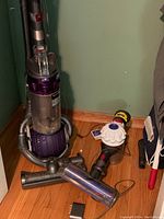 Full view of Ball Animal DC25 upright vacuum, handheld vacuum and attachments on wood floor against wall