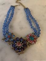 Full necklace laid flat on marble surface showing two blue bead strands and three floral brooch-style centerpieces with purple, blue, pink, and turquoise stones.