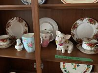 Shelf with Royal Albert Old Country Roses china: teacups, saucers, plates, collectible rose-decorated pig figurines, and coffee mugs.