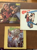 Three Monkees vinyl LP albums laid out on a wood floor showing front covers: 'The Monkees', 'Headquarters', and 'More of The Monkees'. Covers show photographic band images and iconic logo.
