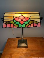 Desk lamp lit showing the stained glass shade with pink, green, and cream colors and floral pattern, metal base with pull chain.