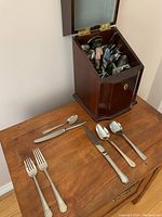 Wooden standing storage box opened, showing assortment of cutlery inside and several pieces laid out on table including forks, knives, spoons.