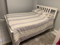 White wooden Ikea bed with vertical slat headboard and paneled footboard, dressed with a striped bedspread and pillows in a small room with tiled floor.