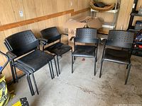 Four black plastic stacking chairs with vinyl seats and armrests arranged in a room with wood paneling; some vinyl seats stained and showing use.