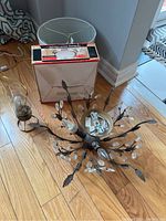 Metal 3-light chandelier with acrylic leaves, ceiling fixture in box, lampshade, and hurricane lamp shown on hardwood floor near wall.