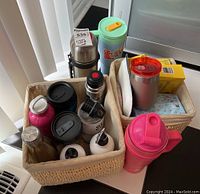 Photo showing two woven baskets containing multiple insulated coffee mugs, water bottles, a thermos, and picnic supplies.