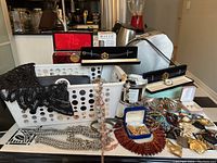 Full lot overview with assorted costume jewelry, watches, and accessories in a white plastic basket on a table with kitchen in background.
