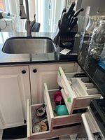 White kitchen cabinetry with three drawers open showing flatware, utensils, scoops and kitchen gadgets, cupboard doors below revealing storage area.