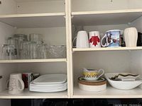 Shelves with a mix of clear glassware, white square plates stacked, assorted mugs including one with a Canadian flag, and ceramic bowls and a floral patterned plate.