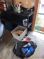 Overall view of shelving unit with spray bottles, a portable heater, a white bin on the floor with paper folders, and two bags near the entrance of a garage or storage area.