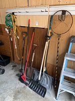 Photo of various garden tools leaning against a wall, including electric hedge trimmer, shovels, rakes, and fishing net along with a small ladder.