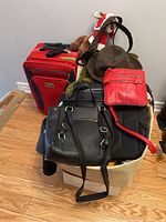 Red carry-on suitcase with black trim and zipper, various hats including felt fedora and woven style, handbags including black backpack and red crossbody, and a stuffed animal on top of a wicker basket.