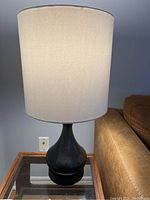 Full lamp with white fabric shade and dark matte ceramic base, standing on glass table.