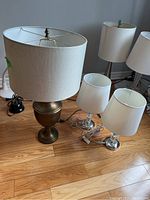 Photo showing all three lamps on wooden floor, two smaller bedside lamps with silver metal bases and one large table lamp with urn-shaped brass base and large fabric shade.