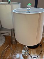 Two lamps with white fabric shades and clear glass cylindrical bases on wood floor. Top finial visible with a green tag.