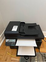 Full front view of black HP LaserJet M1217nfw MFP printer with paper loaded in output tray and scanner lid top visible.