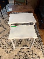 Three nesting tables arranged in descending order by size on a patterned rug. The two smaller tables are painted white with visible wear and uneven paint. The largest table is natural wood with visible watermarks and barley twist legs.