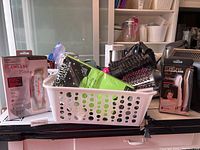 Full view of basket containing multiple electric hair curling brushes, hair curling wand, grooming supplies in packaging and tissue paper, on countertop.