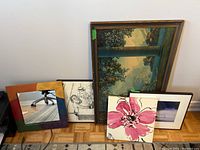 Photo showing all prints and mirror arranged on floor: multi-colored framed mirror, vintage classical Parrish style print, floral print, black and white cartoon, and misty lake signed print.