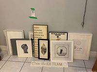 Several framed prints arranged on floor against wall, including typographic and illustrated artworks and a wooden plaque