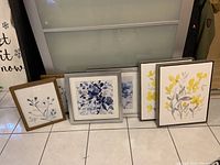 Six floral-themed framed prints displayed side by side on the floor with varying frame colors and sizes.