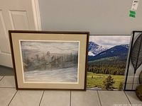 Two prints leaned against a wall. The larger is a giclee canvas print showing a mountain landscape with a field and trees. The smaller one is framed artwork with a misty, ghostly landscape and human-like figures.
