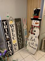 Five wooden door greeter signs standing upright against a wall, including Trick or Treat, two Welcome signs, a pastel bunny, and a snowman with a Let it Snow message