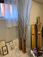 Two tall brown resin vases positioned on the floor against a wall, each filled with long faux wood branches. Also pictured nearby wall art and flowers.