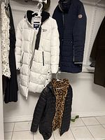 Three winter coats hanging in closet: white Reebok puffer coat, navy quilted coat, black short puffer coat with leopard lining.