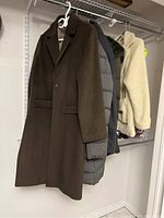 Five winter coats on hangers displayed on wire shelving in basement, including wool, puffer, fleece, and safety vest.