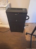 Black air purifier with side handle and caster wheels on hardwood floor next to white dresser and door.