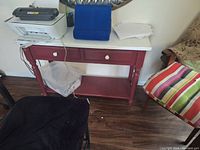 Red wooden desk with drawers, white printer, blue organizer, and papers on top, placed near striped and brown chairs on wood flooring.