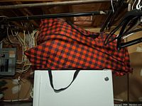 Artificial Christmas tree storage bag with red and black plaid pattern hanging on rack in basement.