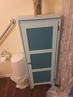 Light blue wooden cabinet with three teal inset panels on door, single silver knob handle, photographed indoors near a trash bin and wall decor.