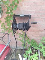 Black outdoor hose coiled on a black metal wall-mounted reel against a brick wall, beside a chair.